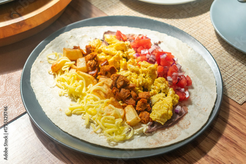 Photograph of an open burrito with egg, cheese, sausage, and onion with tomato placed in a tortilla on a dark plate with natural light with a composition of plates around it.