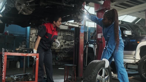 Latin female mechanic worker fixes automotive wheels and talks to a Black coworker who maintains car suspensions in garage station, technician engineering service jobs in vehicle transport industry.