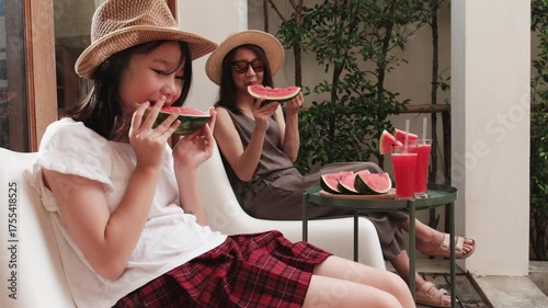 Chilling Asian family holidays, casual girl and mum enjoy eating pieces of fresh red watermelons sliced and smoothie drinks, happiness relaxing on vacation, and hot summertime at outdoor terrace.
