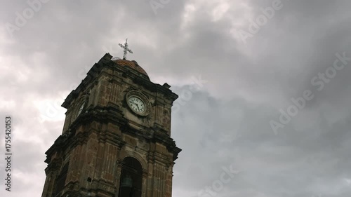The stone clock tower of a historic colonial church stands against a dramatic, overcast sky, as the camera tilts down to reveal the building's full, timeless facade and entrance.