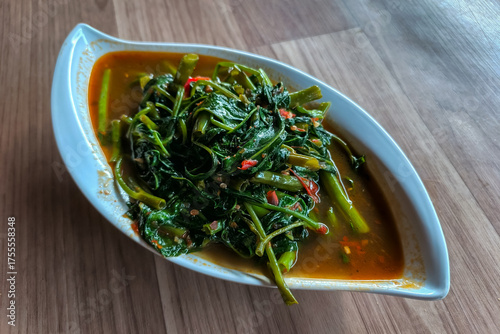 a serving of Kangkung Belacan (stir-fried water spinach with spicy chili shrimp paste) in a white, leaf-shaped bowl on a wooden table.