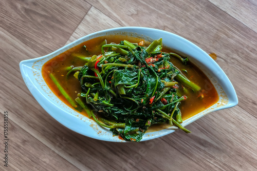 a serving of Kangkung Belacan (stir-fried water spinach with spicy chili shrimp paste) in a white, leaf-shaped bowl on a wooden table.