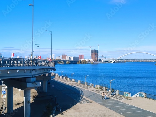 Sunny autumn day on the Dnipro River embankment in Kyiv. View of the river, bridges, and modern urban development on the left bank. Bright blue sky and water.