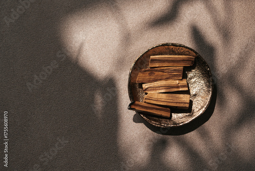 Palo Santo sticks on golden plate, illuminated by artistic shadows of leaves. Combination of organic textures and moody lighting enhances the feeling of tranquility and ritualistic energy. Cropped.