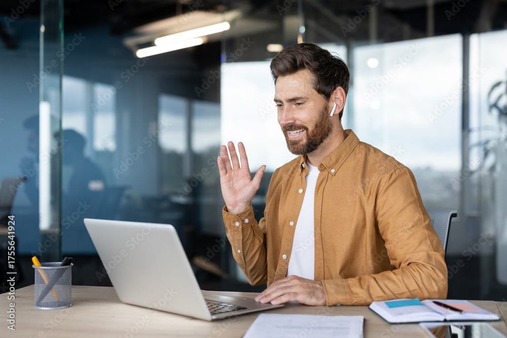 Fototapeta premium Happy bearded man conducting a virtual meeting on his laptop from a modern office, smiling and waving, embracing remote work and digital communication