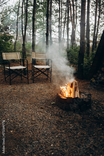 A smoky campfire burning in a small stone fire pit with two folding chairs set up on a gravel clearing in the middle of a dense, dark pine forest, perfect for a weekend escape.
