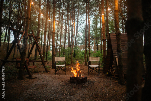 A warm, glowing campfire burning between two canvas folding chairs in a dark, dense pine forest, with string lights illuminating the scene in the evening.