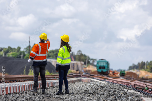 Two railway engineers wearing helmets and reflective jackets inspect new rail tracks under construction.