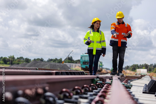 Two engineers wearing safety helmets and high-visibility jackets inspecting a railway track at a construction site. 