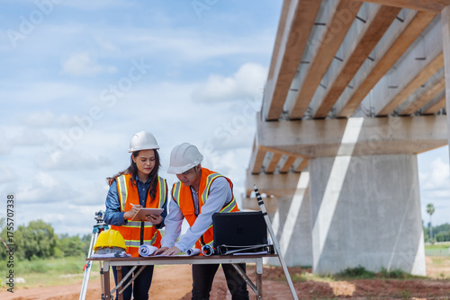 Two civil engineers wearing helmets and safety vests discuss project plans using a tablet at a construction site. 