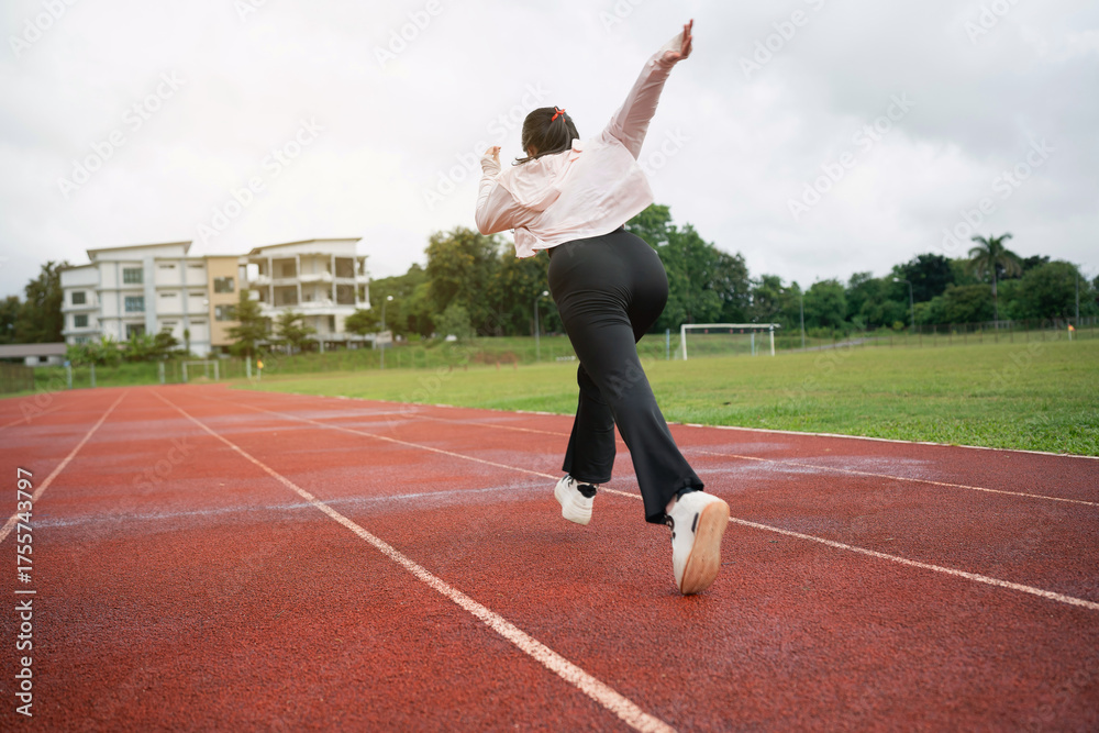 Fototapeta premium Young woman wearing formal attire running on a track field, showcasing determination and energy in an athletic environment, dramatic sky in the background