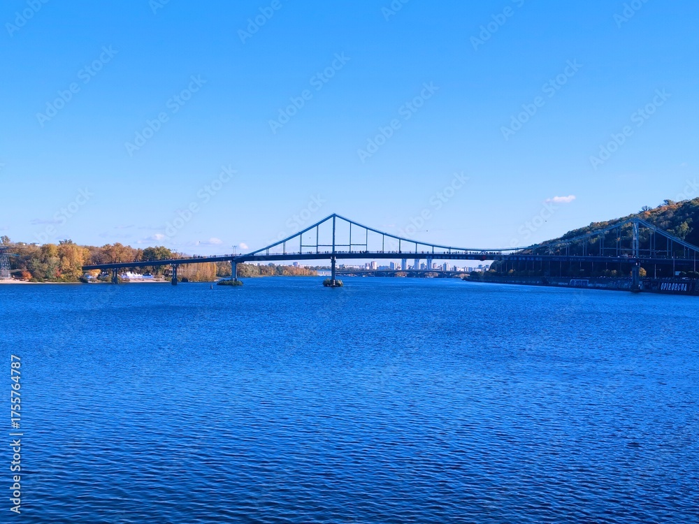 Obraz premium Sunny autumn day on the Dnipro River embankment in Kyiv. View of the river, bridges, and modern urban development on the left bank. Bright blue sky and water.
