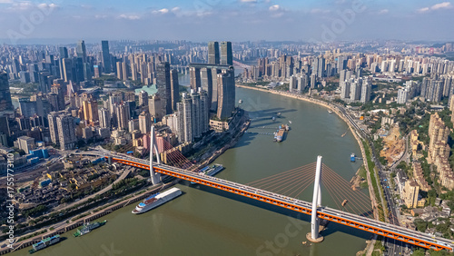A dynamic drone's-eye view of a modern cable-stayed bridge spanning the river in Chongqing, China. Boats and ships navigate the busy waterway below the city's impressive skyline