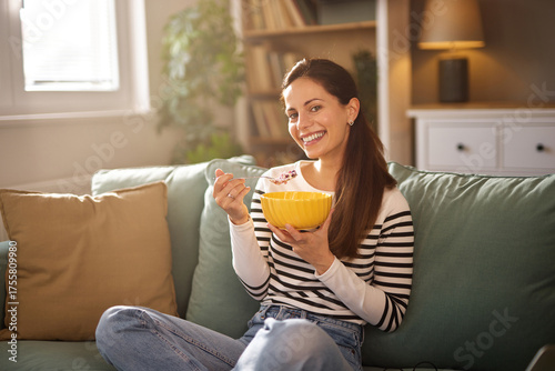 Photos A woman smiles while eating cereal from a bright yellow bowl in a relaxed living room setting