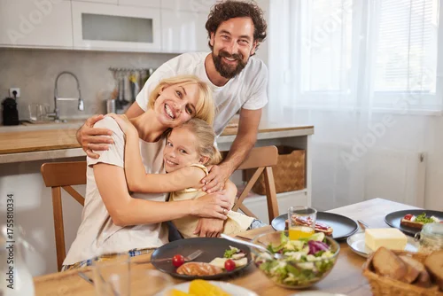 Fototapeta samoprzylepna A family shares a warm moment in their kitchen during lunch. They are smiling and embracing each other, surrounded by a variety of delicious foods on the table.
