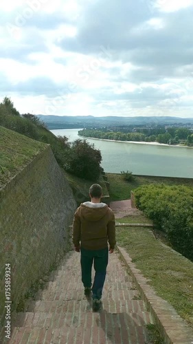 Man descending stairs in Petrovaradin Upper Fortress with view of Danube and Novi Sad. Back view of traveler in motion. Heritage site exploration and cinematic storytelling. Solo tourism moment.