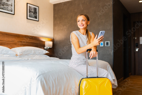 Young woman in hotel room with suitcase sitting on bed looking at phone.