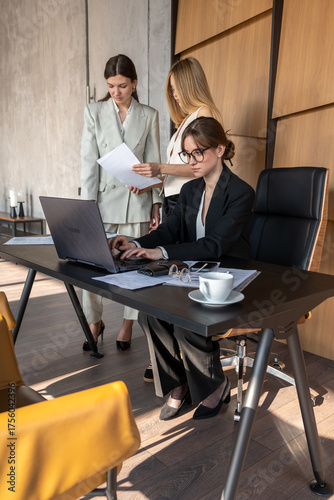 Three professional businesswomen collaborating on a project, one typing on a laptop while two colleagues review documents, working together in a modern office environment