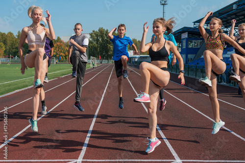 Group of young athletes practicing warm-up and leg exercises using hurdles on an outdoor running track near stadium seating.