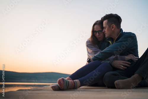 Loving couple sitting close together on a wooden pier by the calm lake at sunset, enjoying peaceful evening moments and connection.