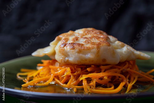 An aesthetic food photograph featuring a close-up of a golden, pan-fried dumpling resting on a bed of finely sliced fresh carrots. The crispy texture of the dough contrasts beautifully with 