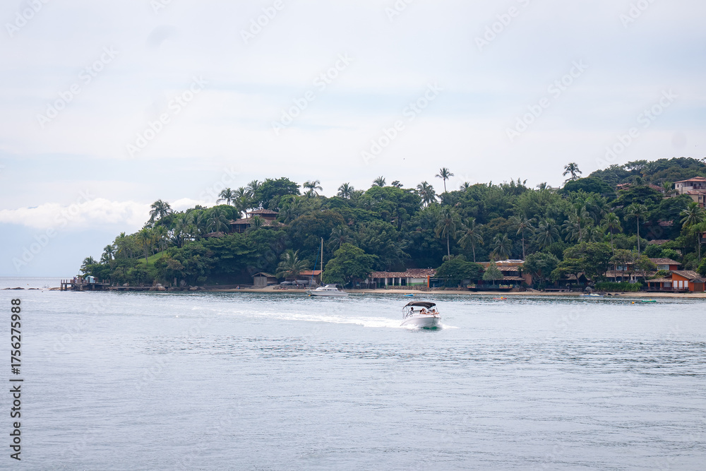 Obraz premium A small motorboat cruises across calm coastal waters, with lush tropical vegetation, and waterfront houses lining the shoreline of the island on an overcast day