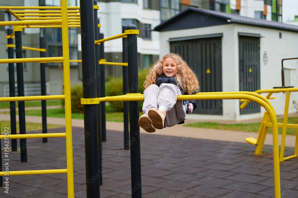 Obraz premium joyful girl playing on monkey bars in urban playground, wearing casual attire. bright yellow play equipment contrasts with surrounding modern architecture, capturing childhood fun
