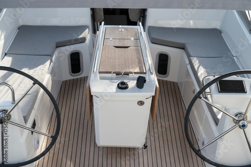 Sailboat with dual steering wheels, comfortable cabins with grey cushions, and wooden decking. Central table with compass and modern design, perfect for a comfortable and luxurious sailing experience.