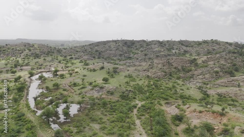 Aerial shot of rocky hills near Masuda, Rajasthan, showing green Aravalli vegetation, small ponds, and water bodies under a cloudy monsoon sky, creating a serene rural landscape.