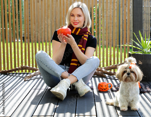 A beautiful blonde girl is sitting on the terrace with a dog, holding a Halloween pumpkin in her hands