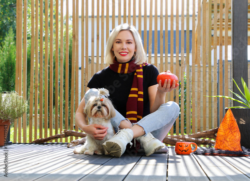 A beautiful blonde girl is sitting on the terrace with a dog, holding a Halloween pumpkin in her hands