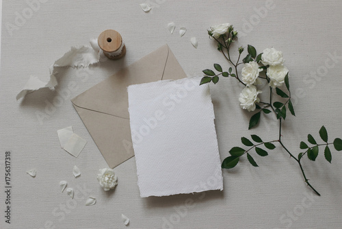 Wedding composition. Feminine mockup scene, white rose flowers, petals, silk ribbon, craft envelope, empty stamps. Blank cotton paper greeting card, invitation. Grey textured background. Flat lay, top