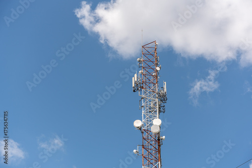 View of a massive telecommunications antenna or transmission tower, supporting various communication antennas for mobile telephony, radio, TV, and internet. The background is a vibrant blue sky.