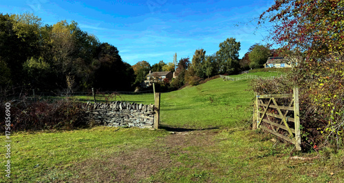 A path leads through a wooden gate towards a farmhouse, surrounded by lush green fields and trees under a bright blue sky. A stone wall adds rustic charm to the scene in Norwood Green, UK