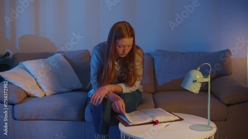 Young Woman Comfortably Sitting on Sofa, Quietly Reading the Holy Bible in Soft Evening Light. Peaceful and Calm Atmosphere for Scripture Study, Spiritual Reflection, Faith, and Personal Devotion.