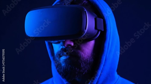 Man wearing a virtual reality headset and hoodie, illuminated by blue and purple lights, looking directly at the camera