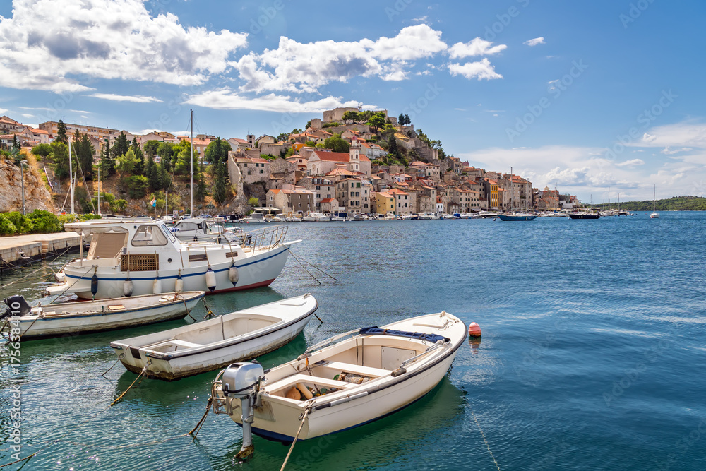 Fototapeta premium Historic European Harbor Scene: Traditional Fishing Boats and Yachts Moored in Crystal Blue Waters, with a Quaint Town and Ancient Fortress on the Hill Under a Summer Sky