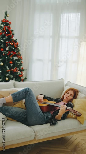 Young woman relaxes on a sofa playing guitar beside a beautifully decorated Christmas tree during the holiday season in a cozy living room
