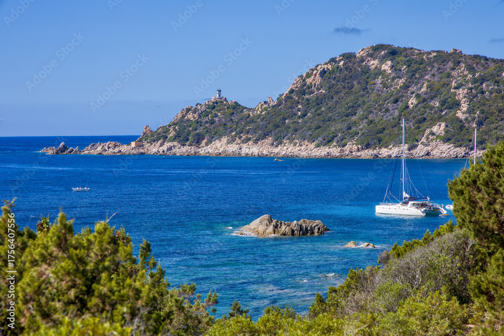 Obraz premium the bay of Cap Muro with its beautiful turquoise water, boats at anchor, and the lighthouse on the hill in sunshine and blue skies