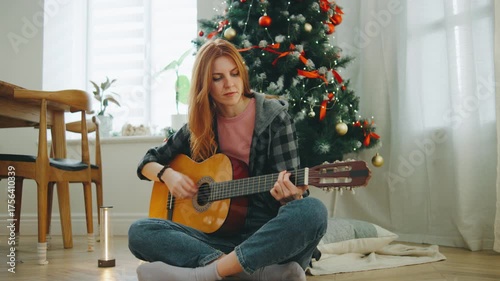 Young woman playing guitar while sitting on the floor next to a decorated Christmas tree in a cozy living room during the holiday season