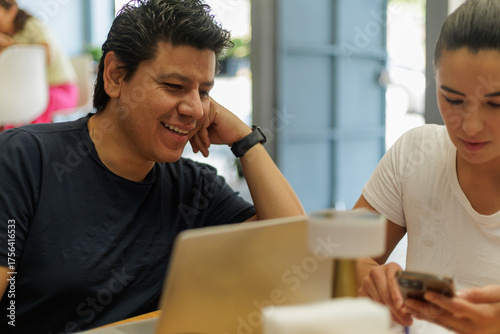 Hombre latino sonriente trabajando en laptop junto a mujer usando smartphone en un espacio moderno.
