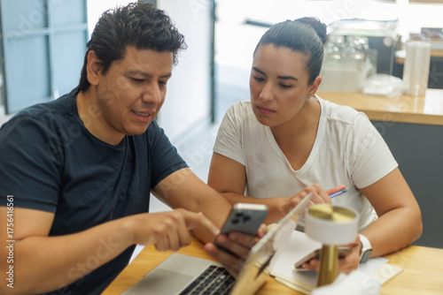 Dos colegas latinos discuten contenido en un smartphone y laptop, colaborando en un espacio de trabajo moderno.
