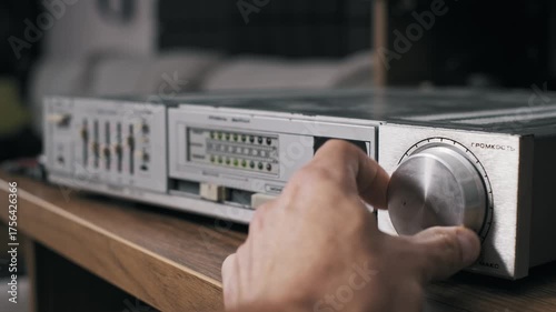 Side angle view of a male hand operating a retro hi-fi amplifier turning the volume knob and pressing buttons. The vintage system has a silver faceplate and green LED indicators on a wood shelf.
