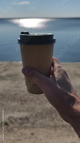Vertical POV shot of a hand holding a brown paper coffee cup with a black lid. Sunny reflection on calm blue water is visible in the soft focus background during the morning.