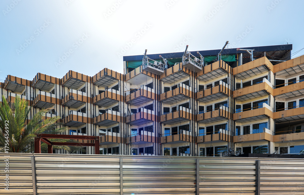 Obraz premium Modern apartment building under renovation with angular balconies, construction equipment on the roof, and a metallic fence in the foreground.