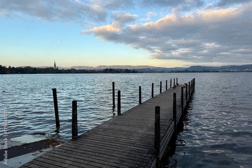 Fototapeta premium Hünenberg am See: A wooden walkway leads out onto Lake Zug, with an autumnal evening atmosphere and clouds in the sky. Symbolic of tranquility, meditation, and peacefulness.