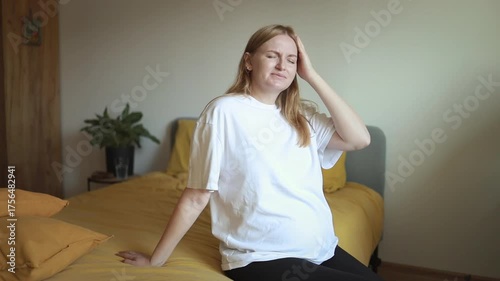 Video of a pregnant woman sitting on bed and touching her head, feeling headache or fatigue. Concept of pregnancy health, stress, pain, motherhood, and prenatal care at home.