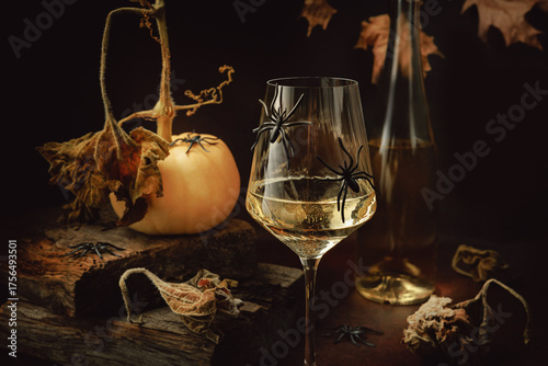Wine glass with white wine and black spiders next to small pumpkin and bottle in dark rustic Halloween composition