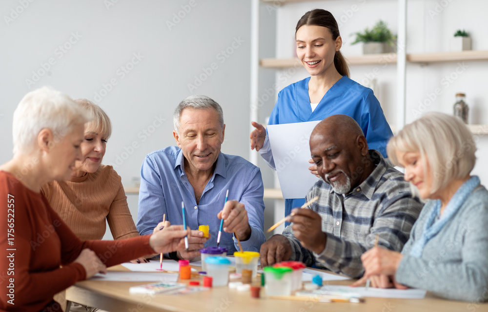 Fototapeta premium Happy multiracial elderly men and women sitting around table and drawing at nursing home, doing arts and crafts together, young lady nurse assisting group of senior people, copy space