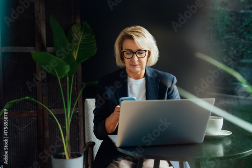 Stylish 50's confident mature businesswoman, middle aged company ceo director, experienced senior female professional, business coach using phone and working on laptop in office. Female leader
